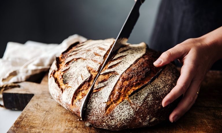 Nahaufnahme von knusprig rundem Laib Brot, das auf einem Holzbrett liegt, gehalten von einer weiblichen Hand, ein Messer teilt es gerade